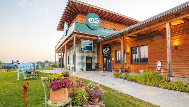 Eis Greissler, © Eis Greissler Gebäude von Eis Greissler mit Holzfassade und dekorativer Kuhfigur im Garten.