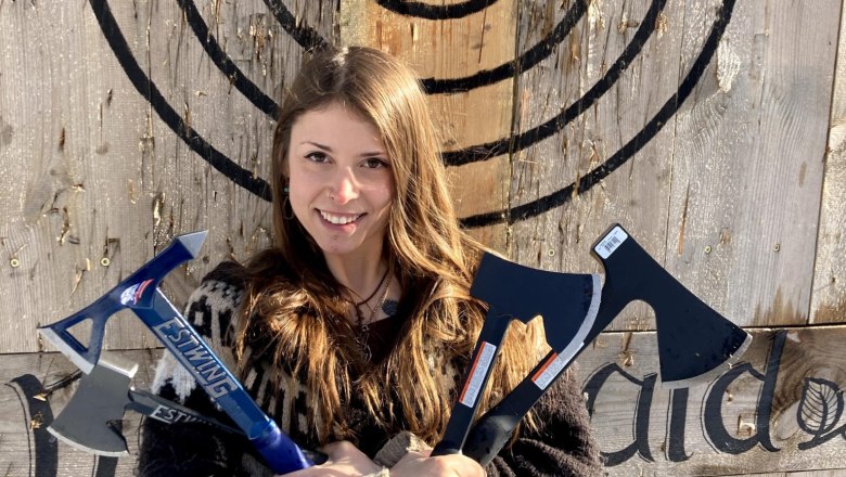 Eine Frau hält drei Äxte vor einer Holzscheibe mit Zielscheibe.