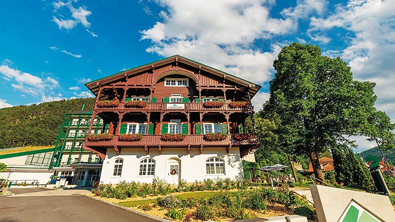 Ein traditionelles, mehrstöckiges Gebäude mit Holzbalkonen und Blumen, umgeben von Bäumen und einem blauen Himmel.