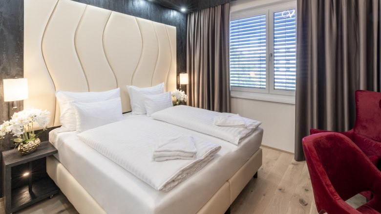 Modernes Hotelzimmer mit Doppelbett, roten Sesseln und großen Fenstern mit Vorhängen.