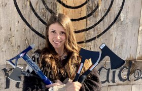 Eine Frau hält drei Äxte vor einer Holzscheibe mit Zielscheibe.