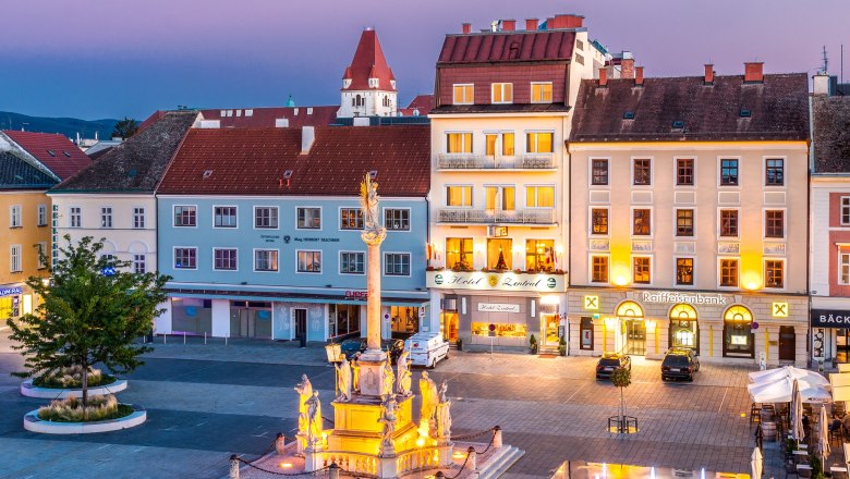 Abendliche Stadtansicht mit beleuchtetem Brunnen und Geb&auml;uden, darunter ein Hotel und eine Bank.