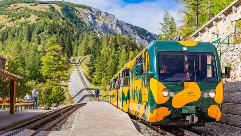 Ein gelb-gr&uuml;ner Salamander-Zug steht an der Station Baumgartner in einer bergigen Landschaft.