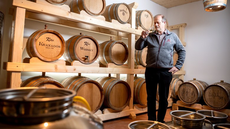 Ein Mann steht in einem Raum mit Holzfässern, die mit 'Kraxados' und 'Whisky' beschriftet sind, und hält ein Glas in der Hand.