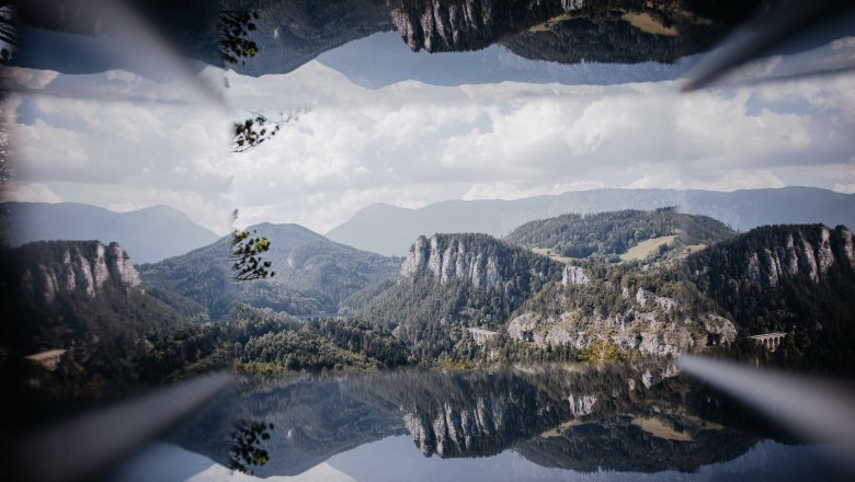 Spiegelbild einer Berglandschaft mit Viadukt und bewaldeten Hügeln unter blauem Himmel.