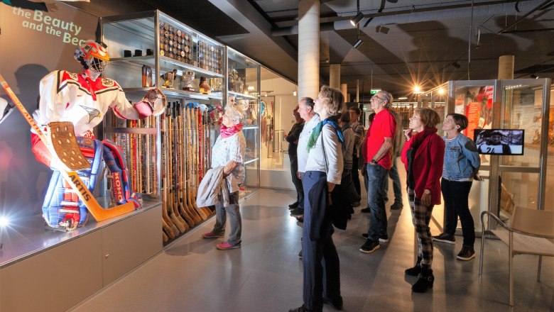 Besucher im Wintersportmuseum betrachten eine Eishockey-Ausstellung mit Ausrüstung und Trophäen.