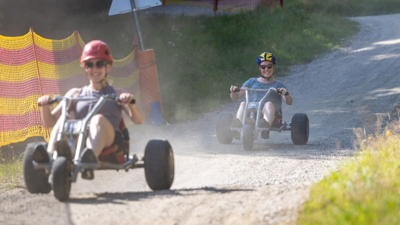 Zwei Personen fahren auf Schotterweg mit Mountaincarts, tragen Helme und Sonnenbrillen.