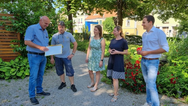 F&uuml;nf Personen stehen auf einem Kiesweg in einem Garten und unterhalten sich. Einer h&auml;lt ein Dokument in der Hand.