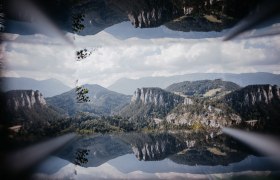 Spiegelbild einer Berglandschaft mit Viadukt und bewaldeten Hügeln unter blauem Himmel.