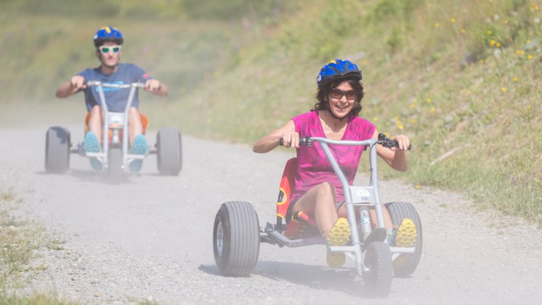 Zwei Personen fahren auf Dreirädern einen staubigen Weg hinunter.